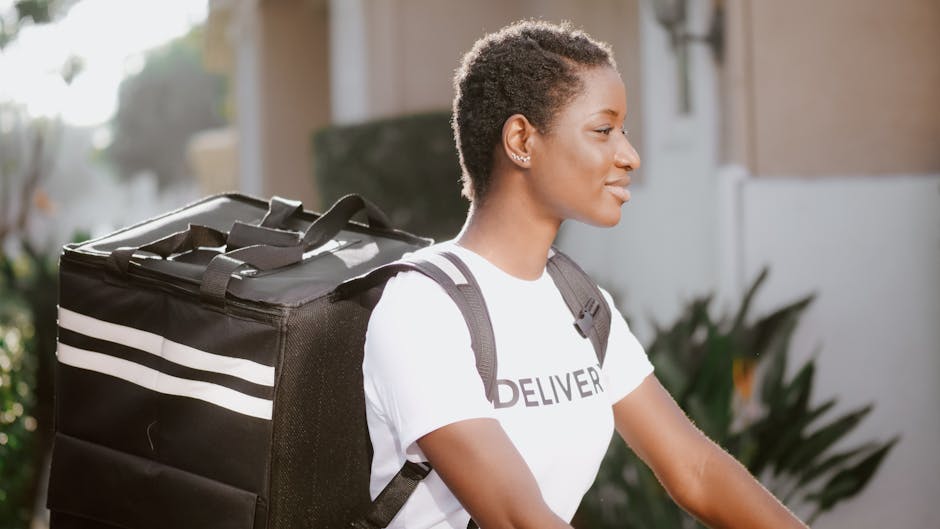 A young woman with short, curly hair and medium skin tone, wearing a white t-shirt with 'DELIVER' printed on it, smiles softly as she rides a bicycle in a suburban outdoor setting during daytime. She